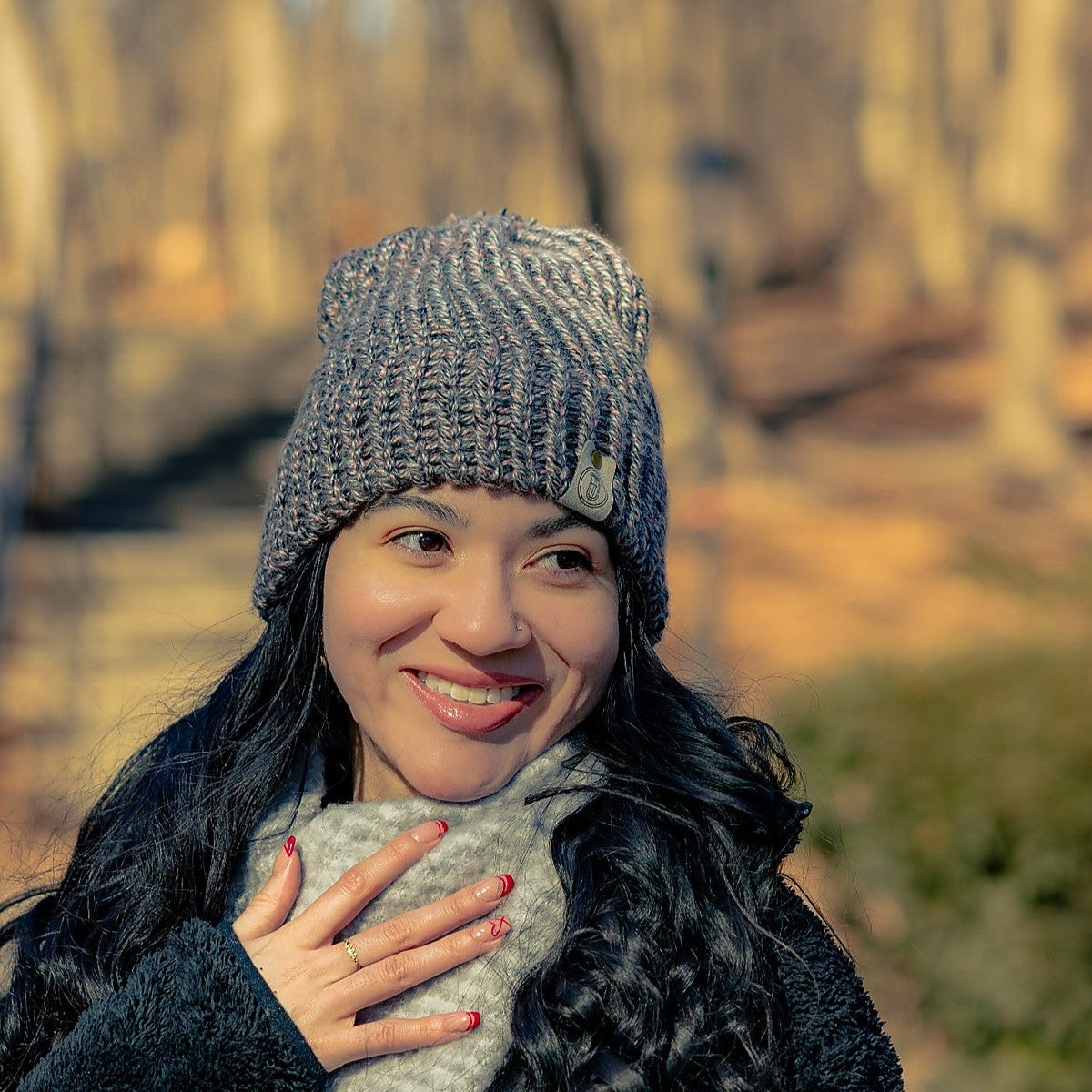 Woman smiling in aHandmade Ash Rose Knit Beanie, with a gray scarf and long curls, standing outdoors in a wooded winter setting.