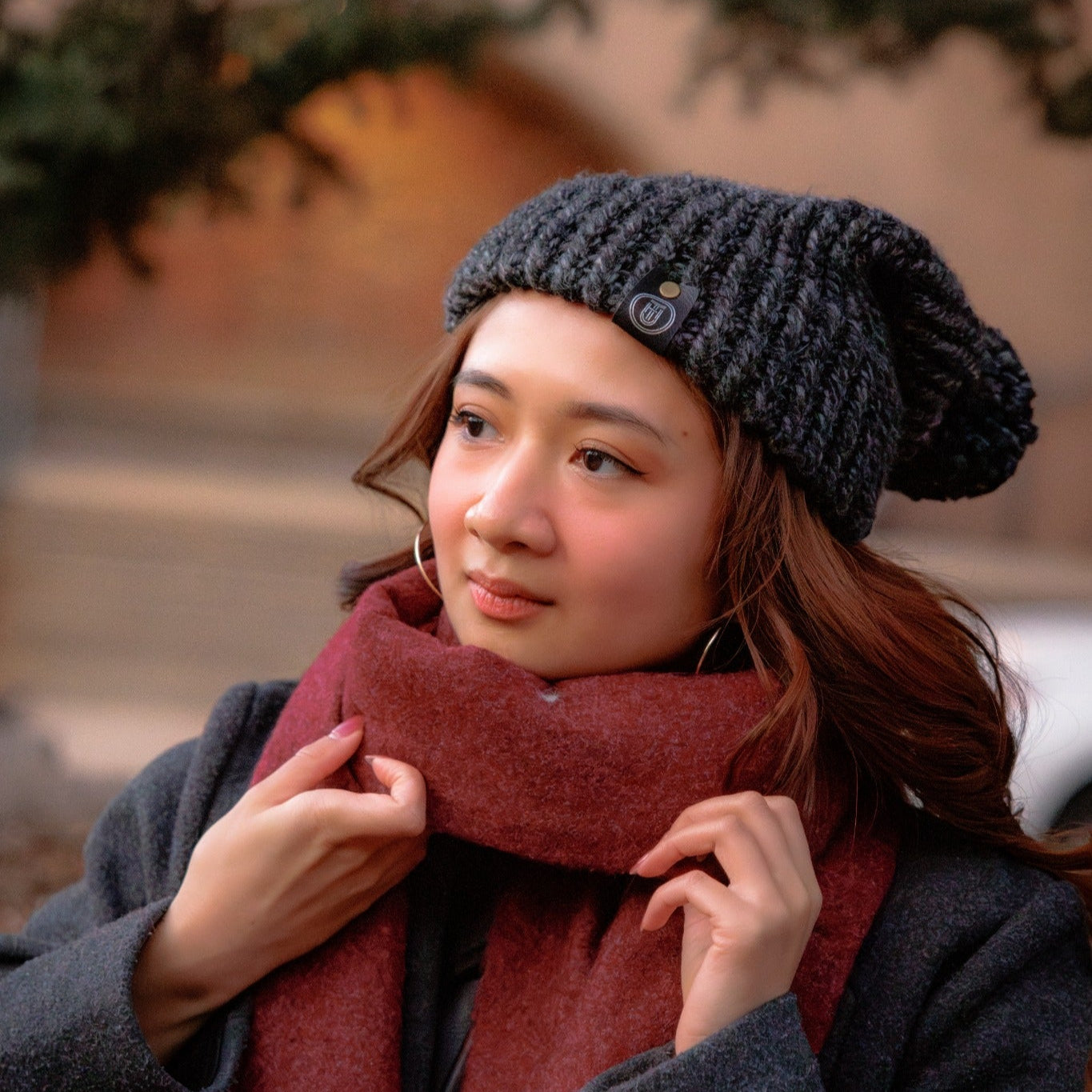 Woman adjusting a Handmade Charcoal Knit Beanie standing outdoors in a gray coat with soft winter lighting.