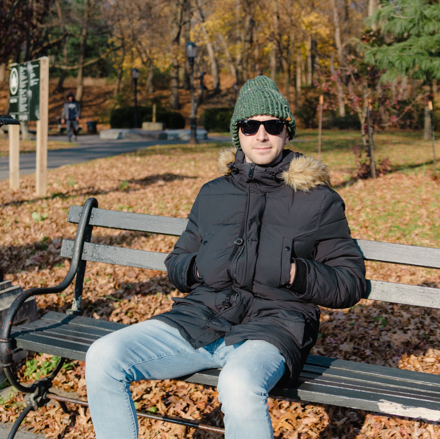Man wearing a Handmade Green Knit Beanie sitting on a park bench in autumn leaves with sunglasses on.
