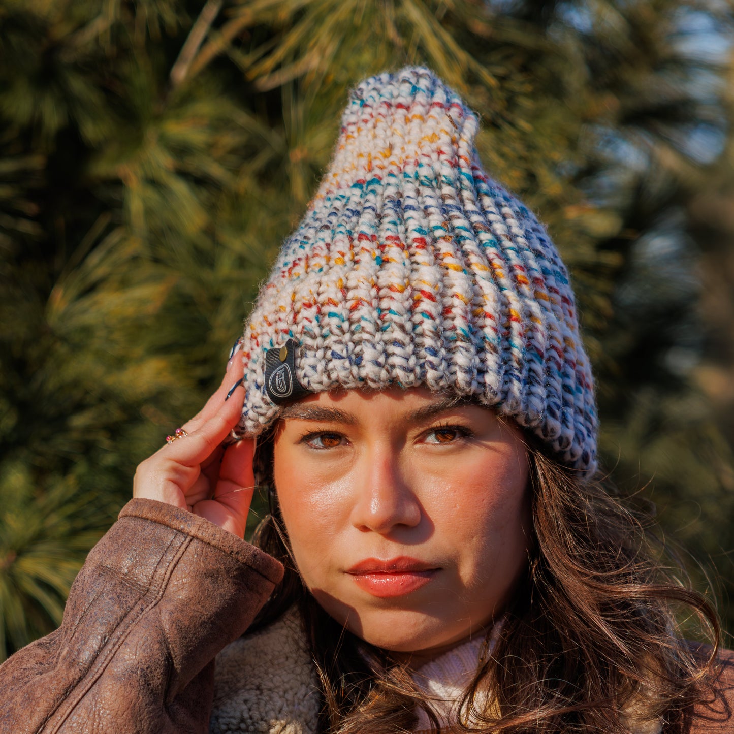 Woman wearing a Handmade Multicolor Speckled Knit Beanie standing in front of a pine tree on a sunny winter day.