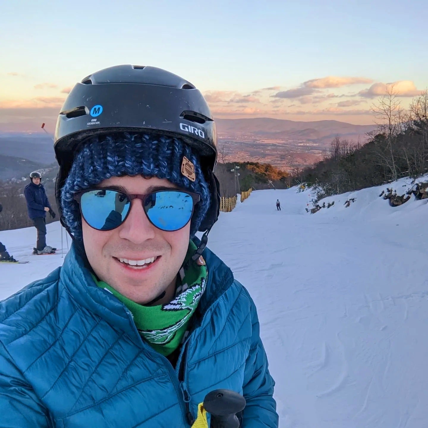 Man wearing a navy blue handmade crochet beanie under a ski helmet, smiling on a snowy mountain slope at sunset.