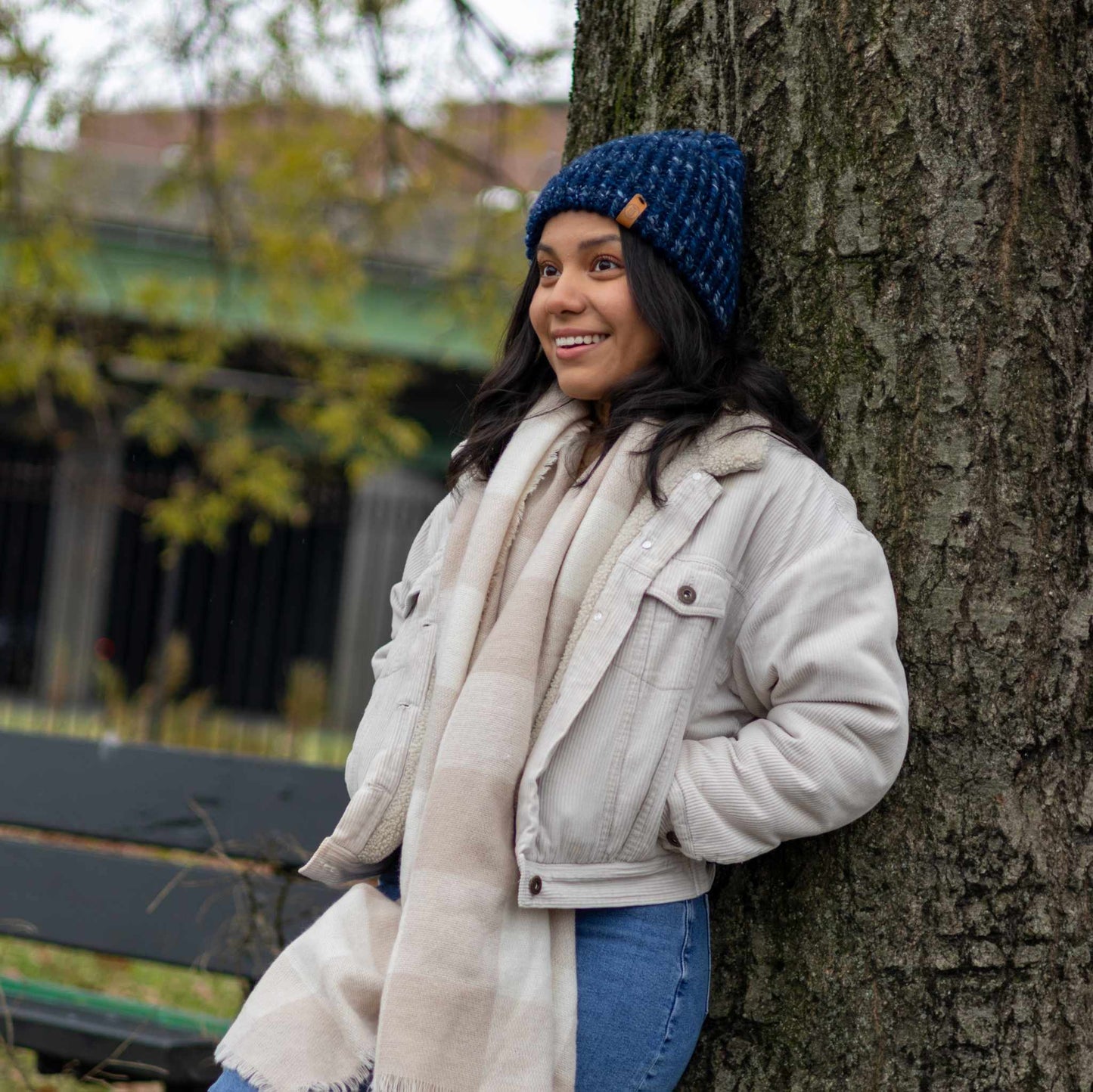 Woman wearing a Handmade Navy Knit Beanie, leaning against a tree in a winter jacket and scarf, smiling outdoors.
