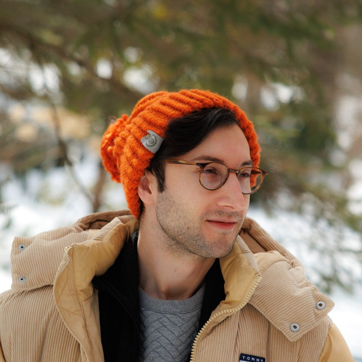 Man wearing a Handmade Orange Knit Beanie with a metal tag, standing outside in a snowy forest wearing a tan winter jacket.