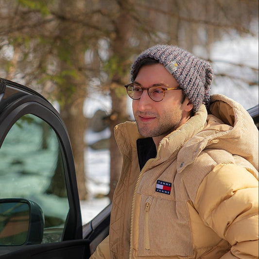 Man wearing a handmade ash rose knit beanie and beige puffer jacket, standing by an open car door with a snowy forest in the background.