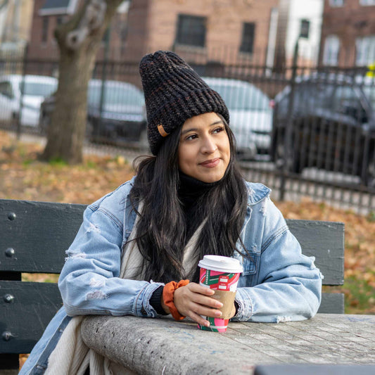 Woman wearing a black and brown handmade crochet beanie, sitting on a park bench in a denim jacket and holding a holiday coffee cup.