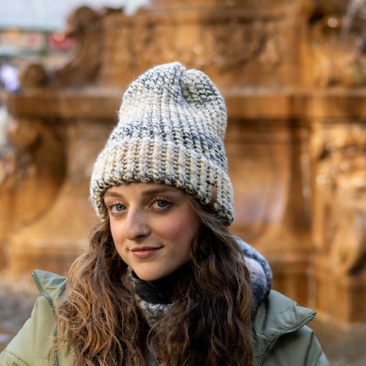 Woman wearing a Handmade Cream & Gray Knit Beanie in front of a golden fountain, bundled in a scarf and winter coat.
