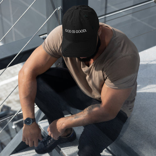 Front view of black Christian GOD IS GOOD Dad Hat worn by seated model in tan shirt