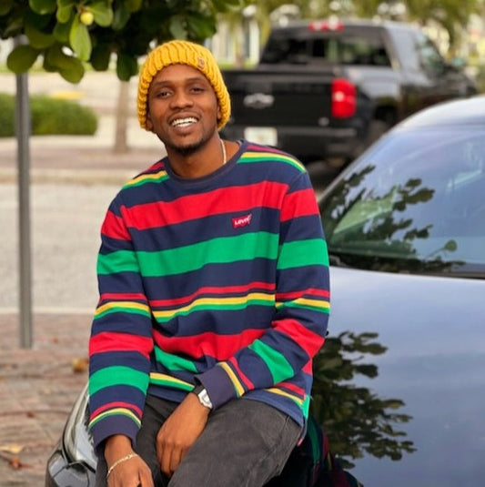 Man wearing a Handmade Sunrise Gold Knit Beanie, smiling outdoors in a striped sweater while sitting on a car hood.
