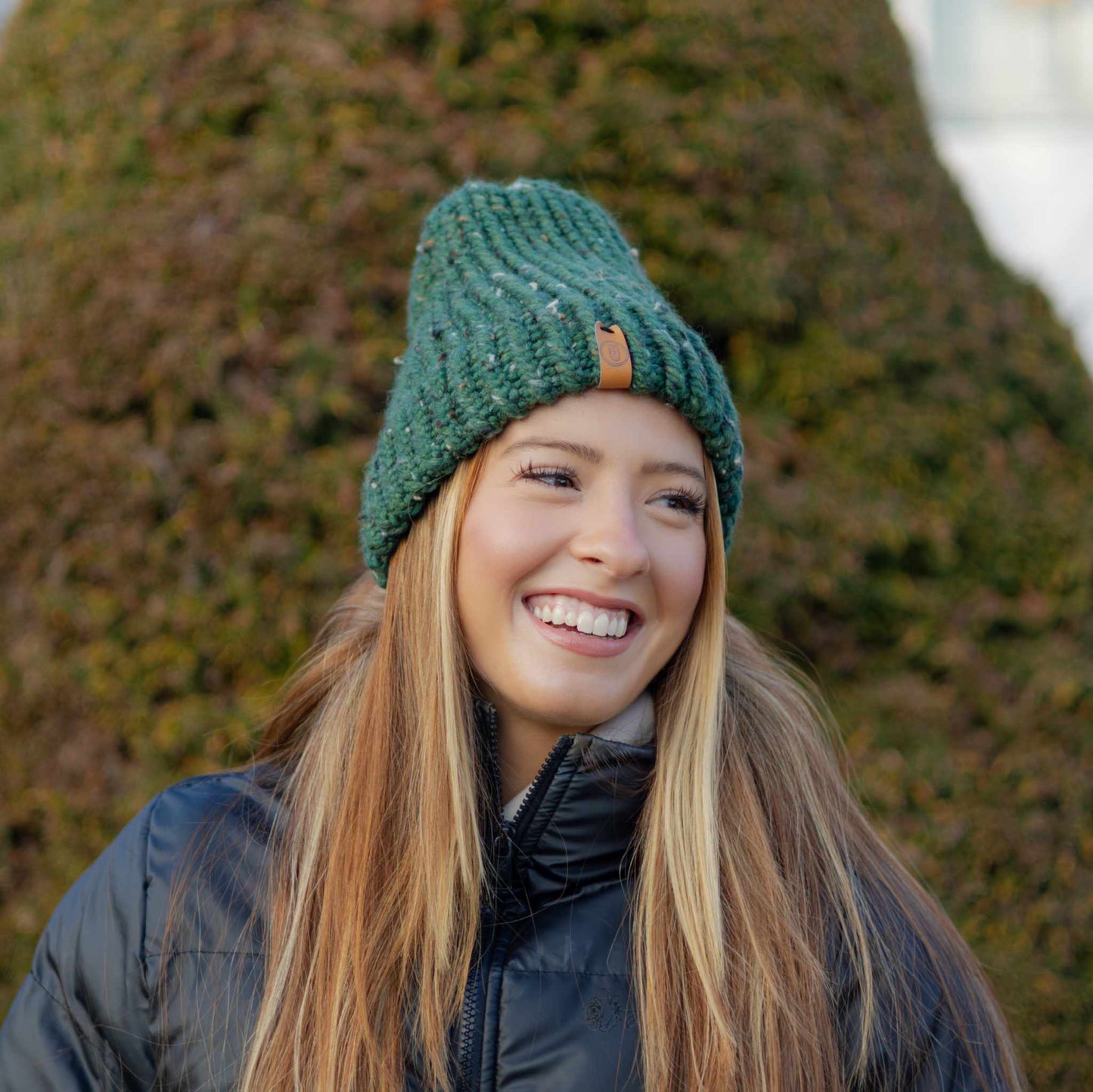 Woman wearing a Handmade Green Knit Beanie, smiling outdoors in a black puffer jacket in front of green hedges.