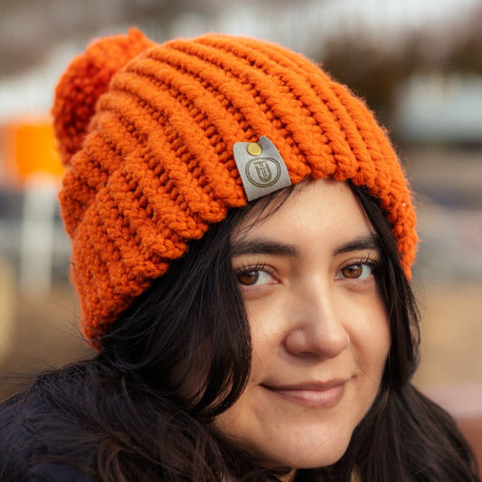 Woman wearing a bright orange handmade knit beanie with a felt tag, smiling outdoors on a winter day.