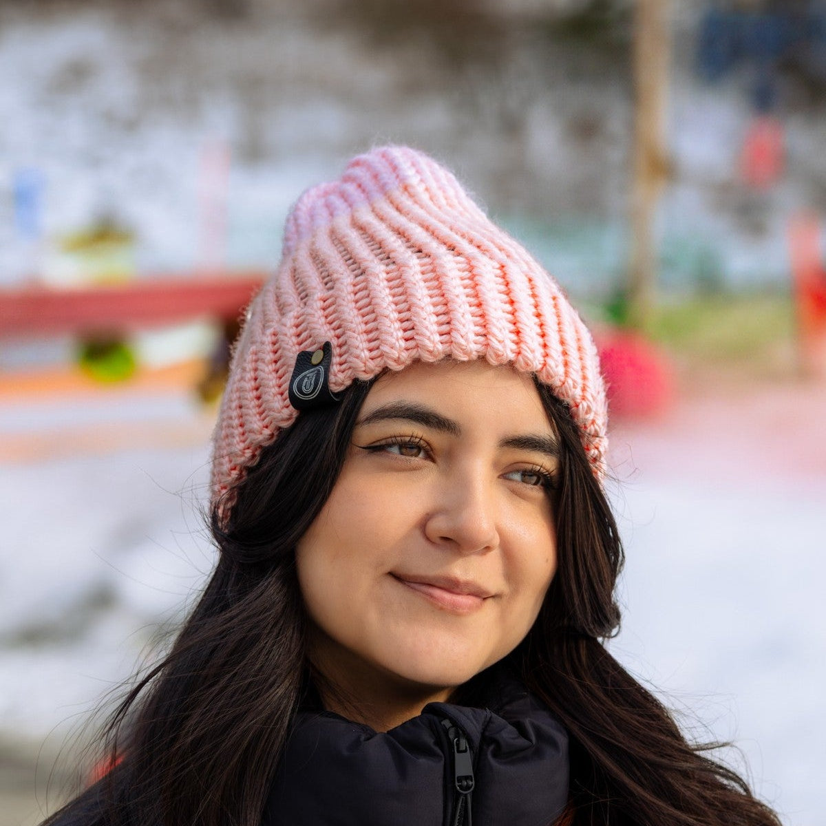 Woman wearing a peach-pink handmade knit beanie and a black puffer jacket, smiling outdoors on a snowy day.